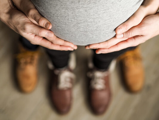 Blick von oben auf einen Babybauch, der von schwangerer Frau und ihrer Partnerin gehalten wird.