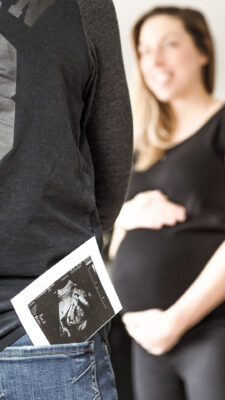 Ultraschallbild steckt in Gesäßtasche der Partnerin, im Hintergrund glücklich strahlende schwangere Frau.