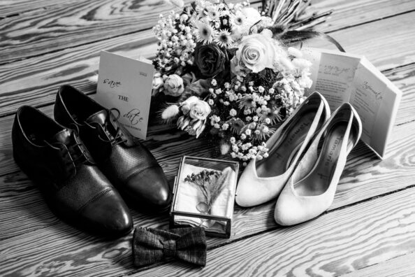 Brautstrauß, Schuhe, Ringe und Einladungskarten auf einem Holzboden in Szene gesetzt.